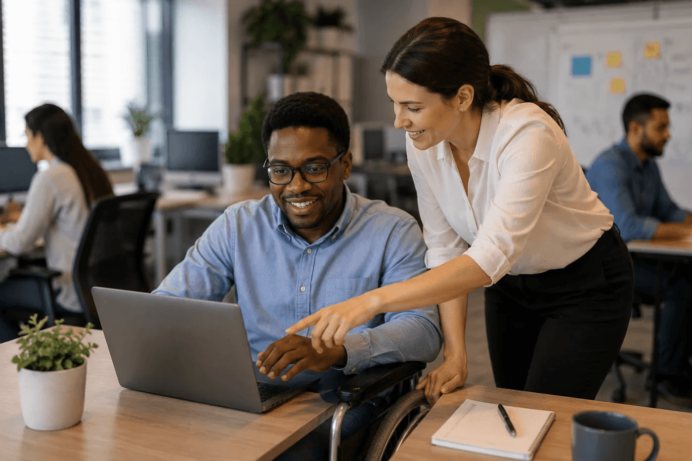 Two colleagues collaborate at a laptop in a bright office; one team member uses a wheelchair.